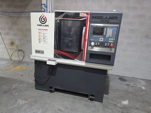 Torno CNC tipo gang de cabina compacta ya en nave, sobre pared de bloque gris: frontal blanco con logo Ingelaser.