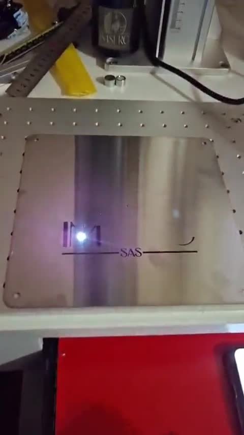 Marcadora láser de sobremesa grabando un logotipo (una 'M' con la palabra SAS bajo una línea) sobre una placa de acero apoyada.